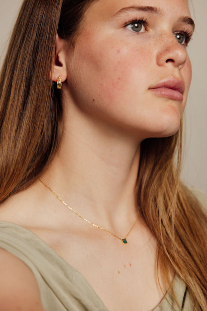 Close-up of a woman wearing a gold necklace with a green gemstone and gold earrings on a neutral background
