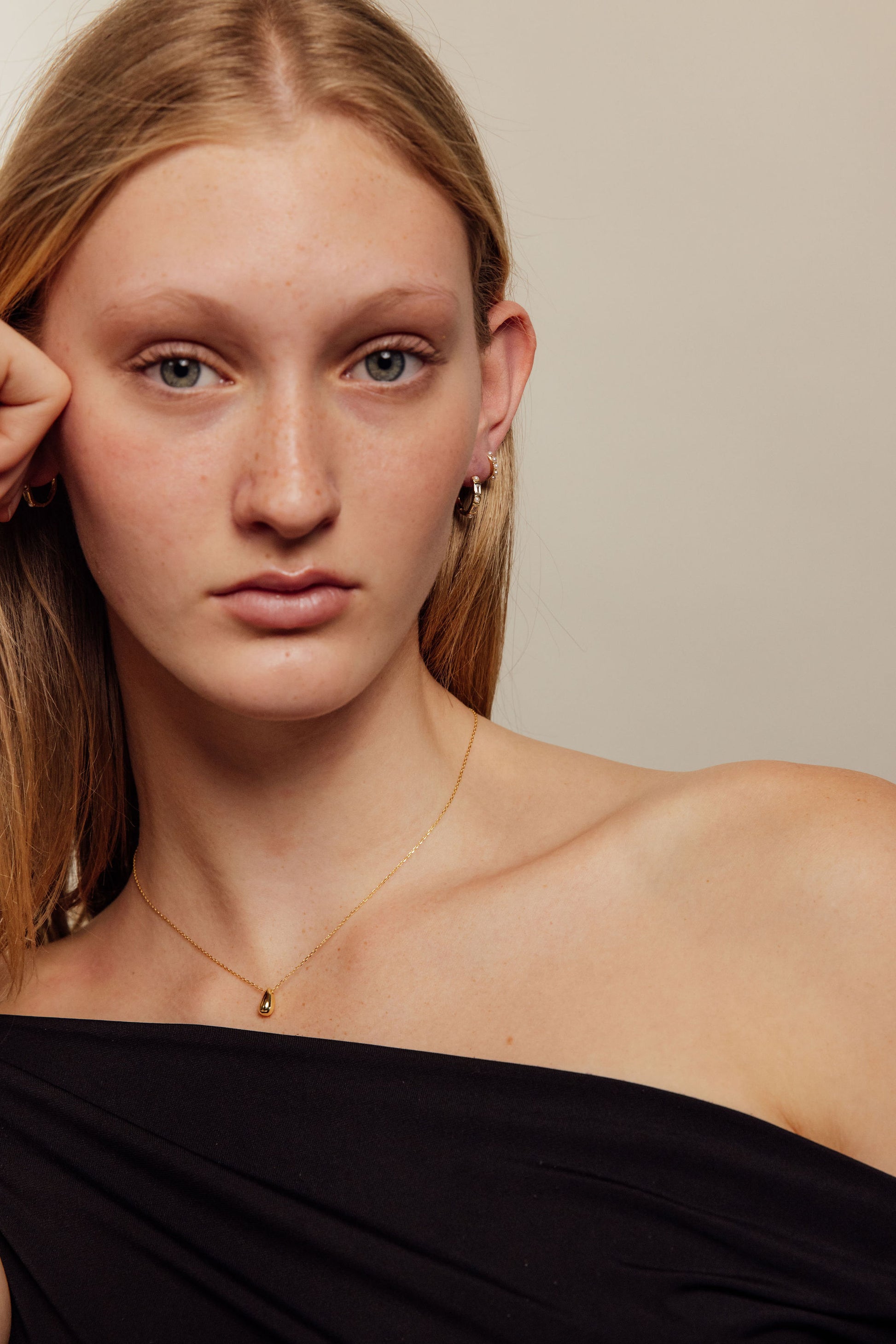 Woman wearing gold earrings and a gold necklace wearing a black off-shoulder top against a neutral background