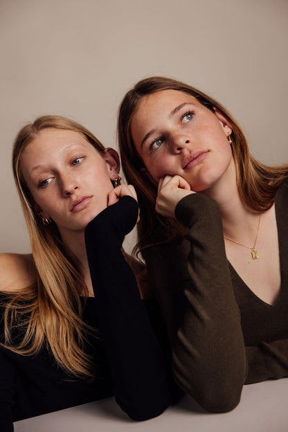 Two women sitting close together with a neutral background, both wearing gold jewellery