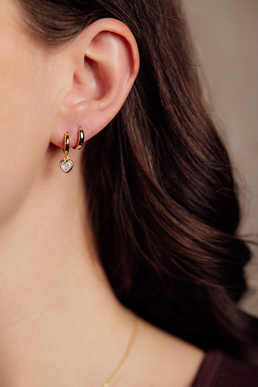 Close-up of a person wearing gold hoop earrings with heart charms.