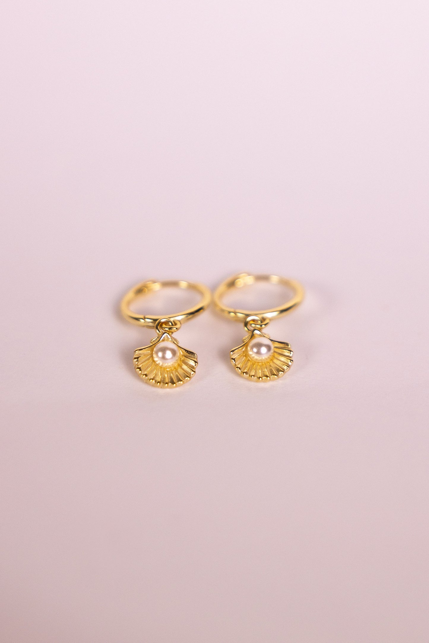 Gold earrings with shell shaped charms featuring a pearl on a pink background