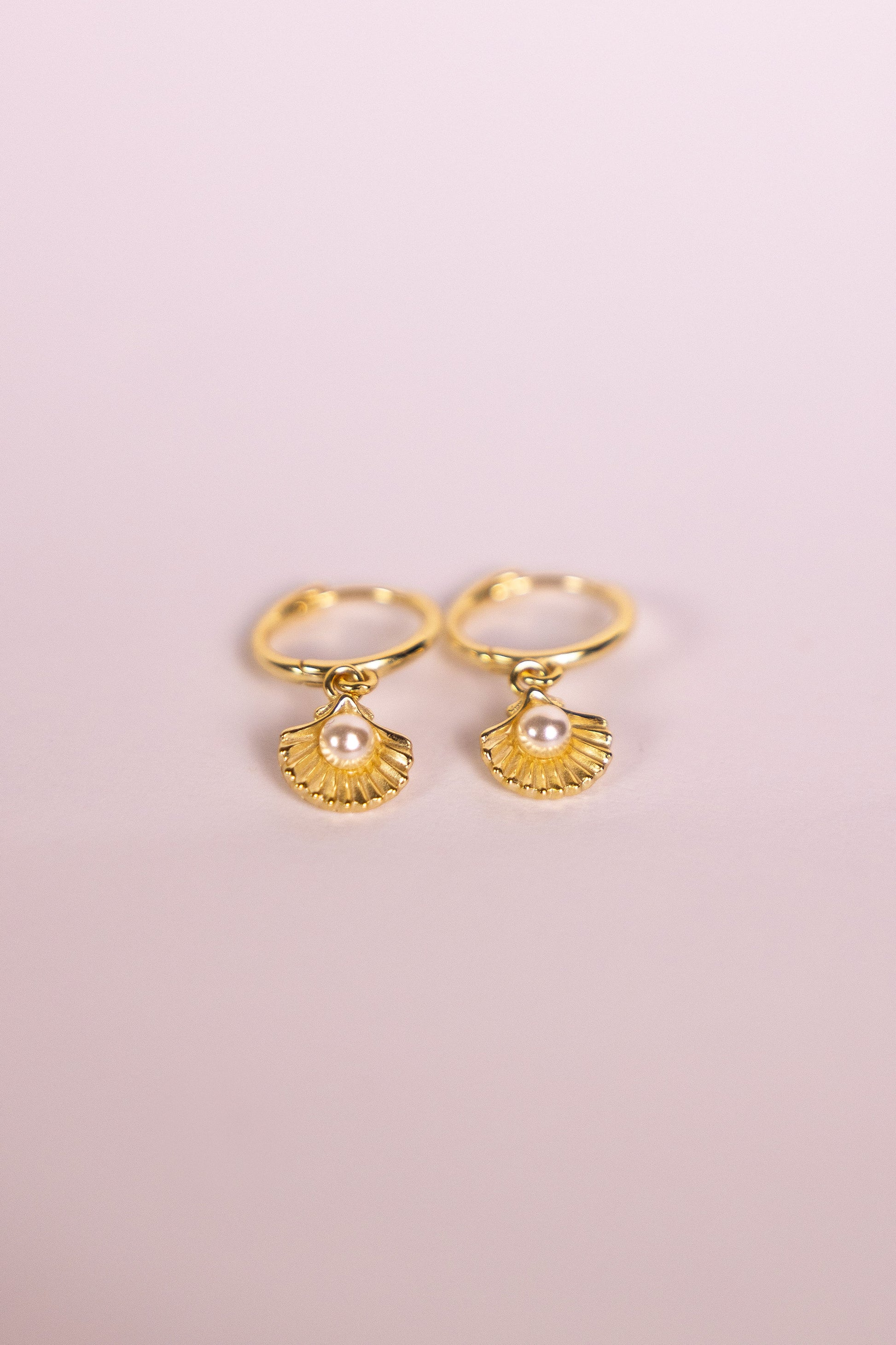 Gold earrings with shell shaped charms featuring a pearl on a pink background