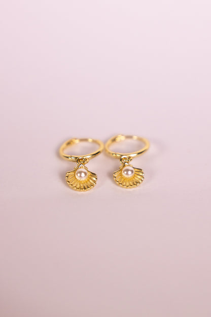 Gold earrings with shell shaped charms featuring a pearl on a pink background