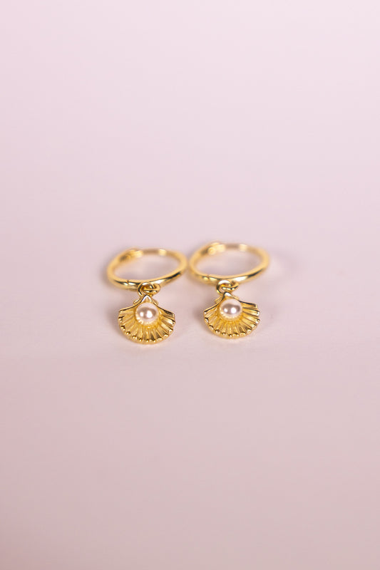 Gold earrings with shell shaped charms featuring a pearl on a pink background