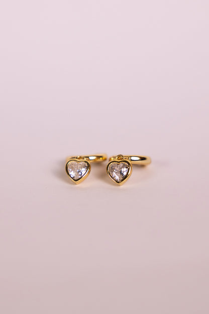 Gold earrings with a heart-shaped charm on a neutral background