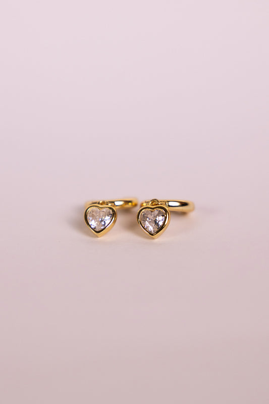 Gold earrings with a heart-shaped charm on a neutral background